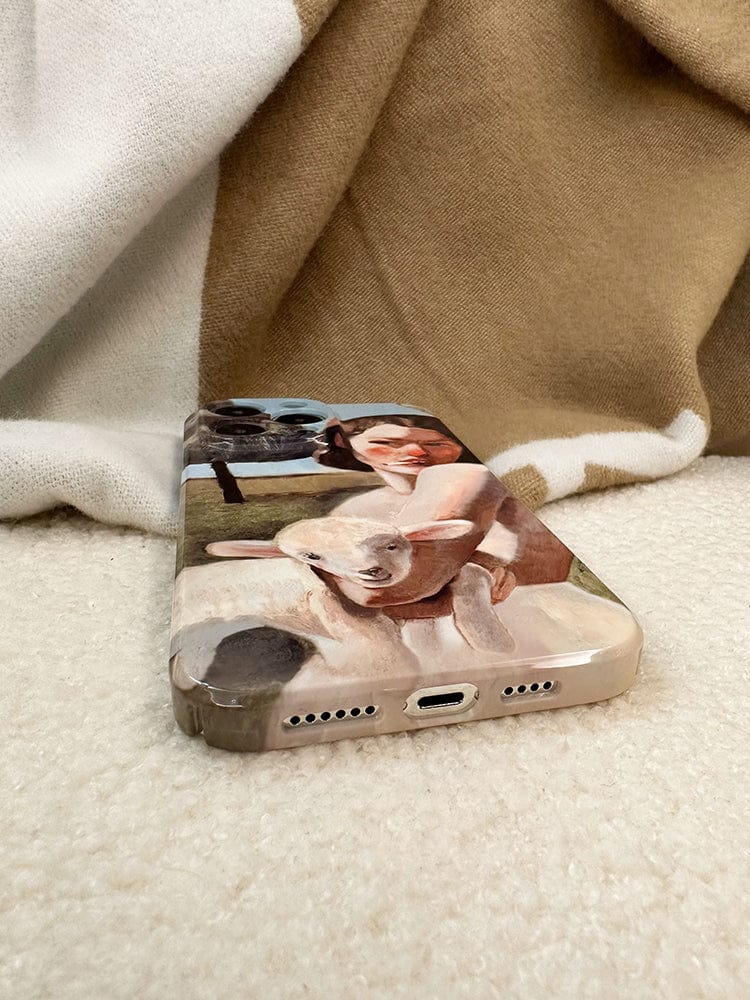 A smartphone rests on a soft, beige blanket. Its Pastoral Art iPhone 16 Pro Max Case features a painting of a woman hugging a white sheep, with a green field and another sheep in the background. The bottom of the phone, showcasing the charging port and speaker holes, is in sharp focus.