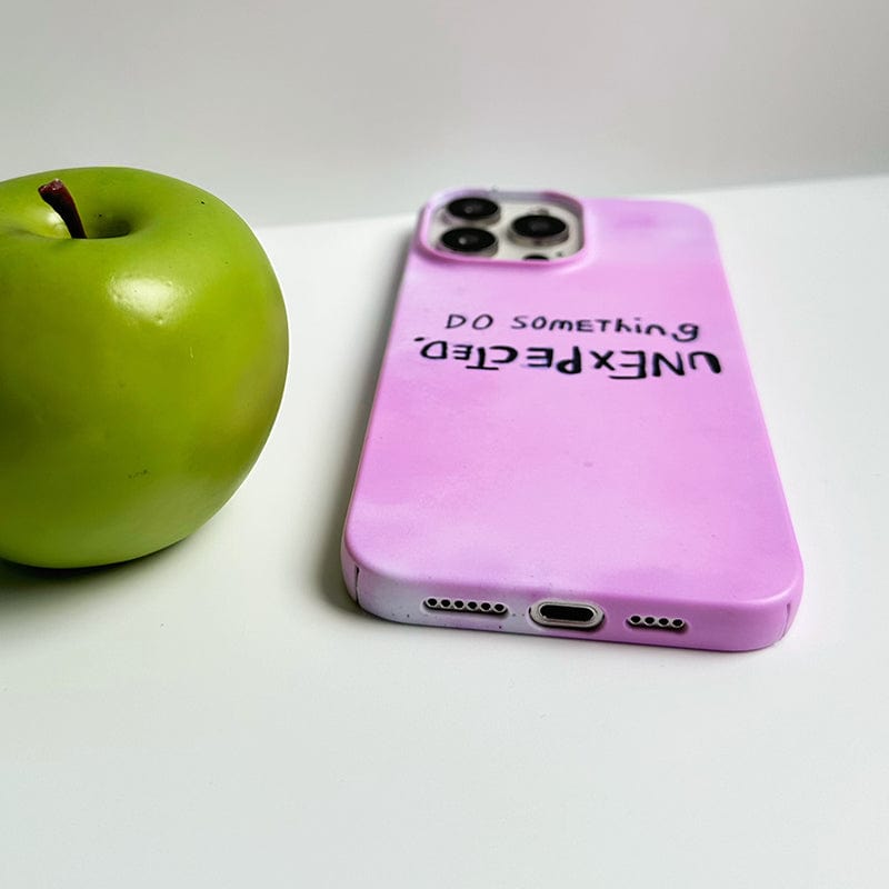 A green apple is placed next to a smartphone. The phone, an iPhone 16 Pro Max, is lying face-down with an Inspirational Pink iPhone 16 Pro Max Case featuring the quote "DO SOMETHING UNEXPECTED" in black letters, where "UNEXPECTED" is inverted. The background is plain white.