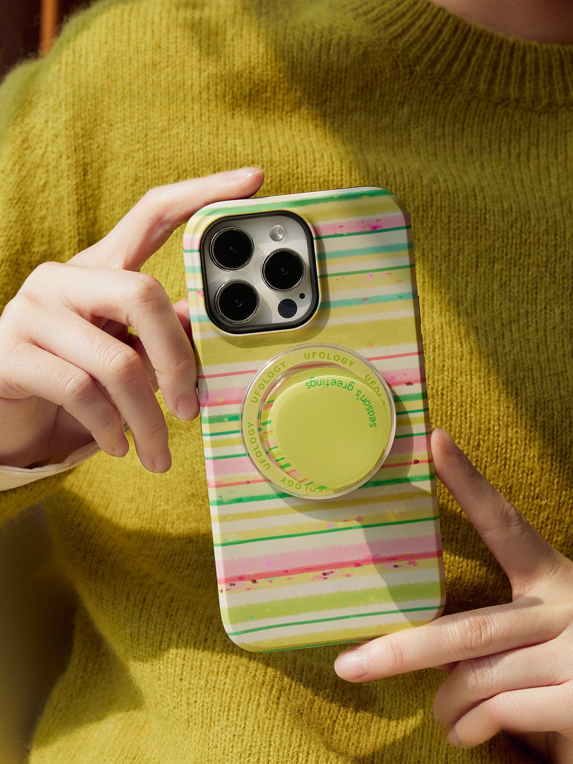 A person wearing a yellow sweater holds an iPhone 16 Pro Max with the Festive Stripes MagSafe Case. This cheerful holiday design features horizontal stripes in green, pink, yellow, and white, and includes a circular grip in the center with the text "UFOLOGY".