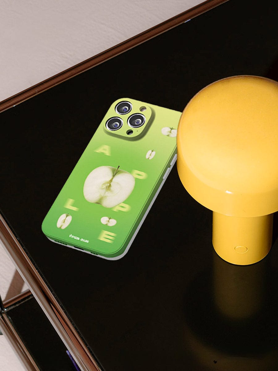 An iPhone 16 Pro Max with a Green Apple Fresh case, showcasing a vibrant gradient design and playful apple-themed images, rests on a reflective dark surface next to a yellow cylindrical object resembling a lamp.