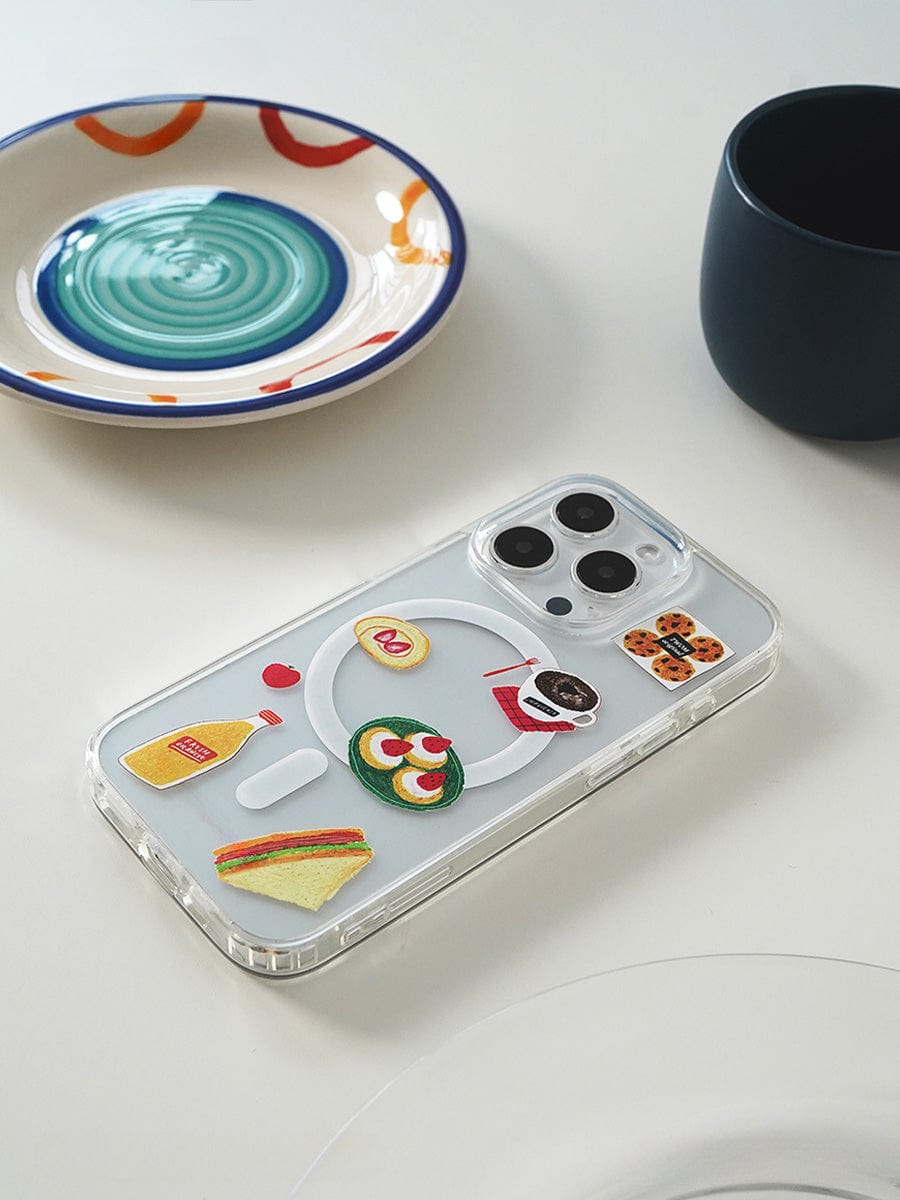 A smartphone encased in a Brunch Time MagSafe iPhone 16 Pro Max Case, featuring a cute 3D food design with stickers of a sandwich, sunny-side-up eggs, a bottle of sauce, and a cup of coffee, sits on a table near colorful ceramic dishes—one with a spiral pattern—and alongside a dark cup.