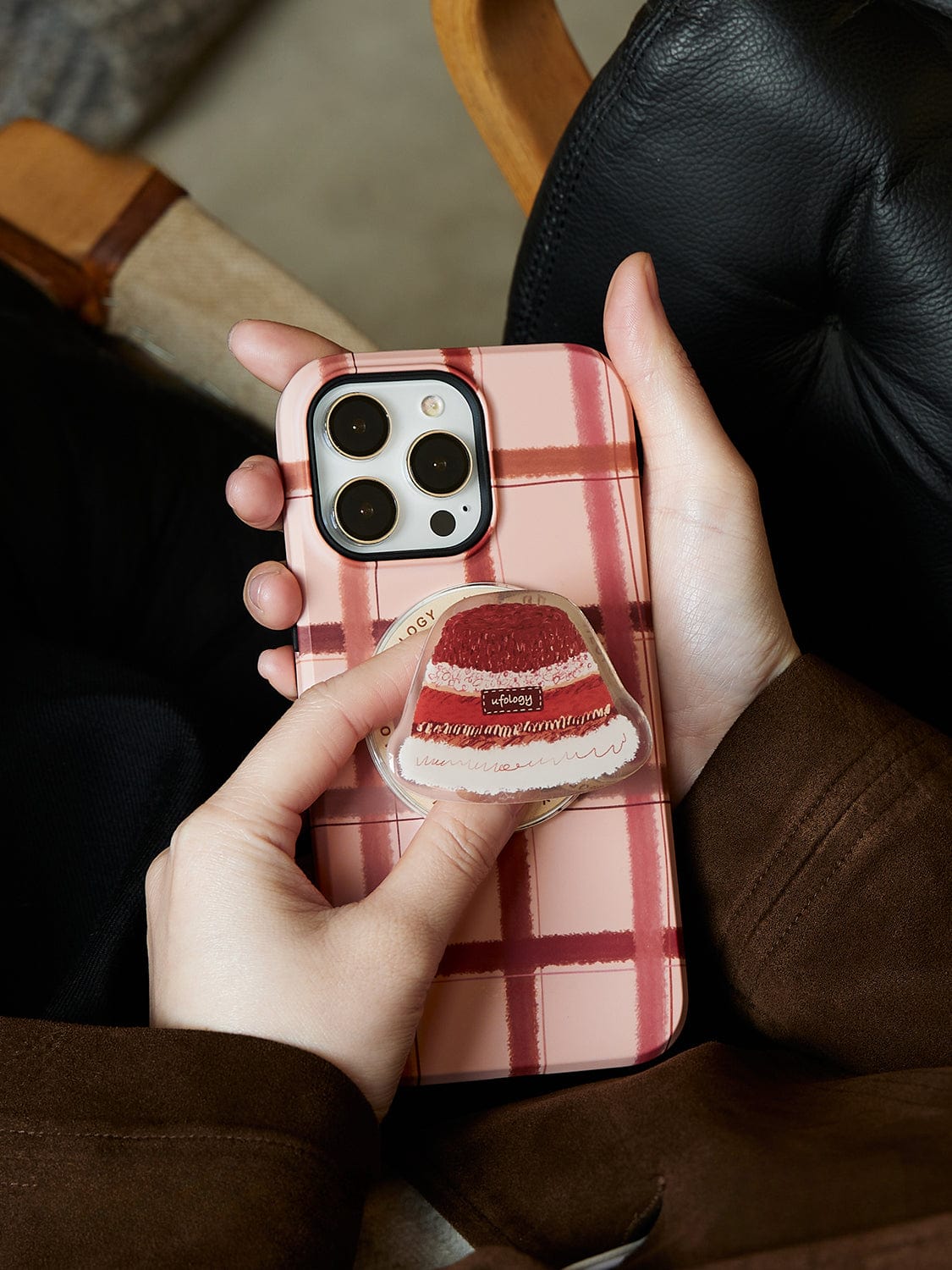 A person holds a Cozy Plaid MagSafe iPhone 16 Pro Max Case | Warm Winter Hat Design with Stand in one hand, while their other hand grips a pop socket attached to the case.