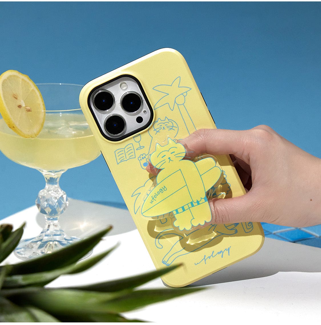 A hand holds the Tropical Cat MagSafe iPhone 16 Pro Max Case, which features a relaxed cartoon cat lounging with a drink under a palm tree, near a swimming ring. A lemon-topped drink sits in the background, and there's a small green succulent in the foreground. The sky is bright blue.