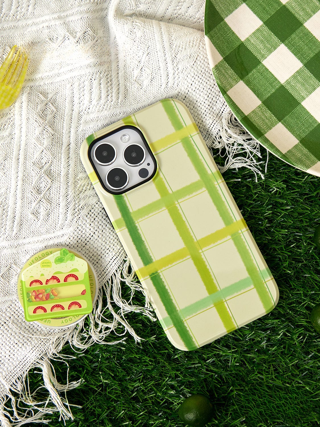 A smartphone encased in the Fresh Picnic MagSafe iPhone 16 Pro Max Case with a green plaid design, a ceramic coaster featuring a slice of fruit cake, a green and white checkered plate, and a fork on a white textured cloth set on grassy surface.