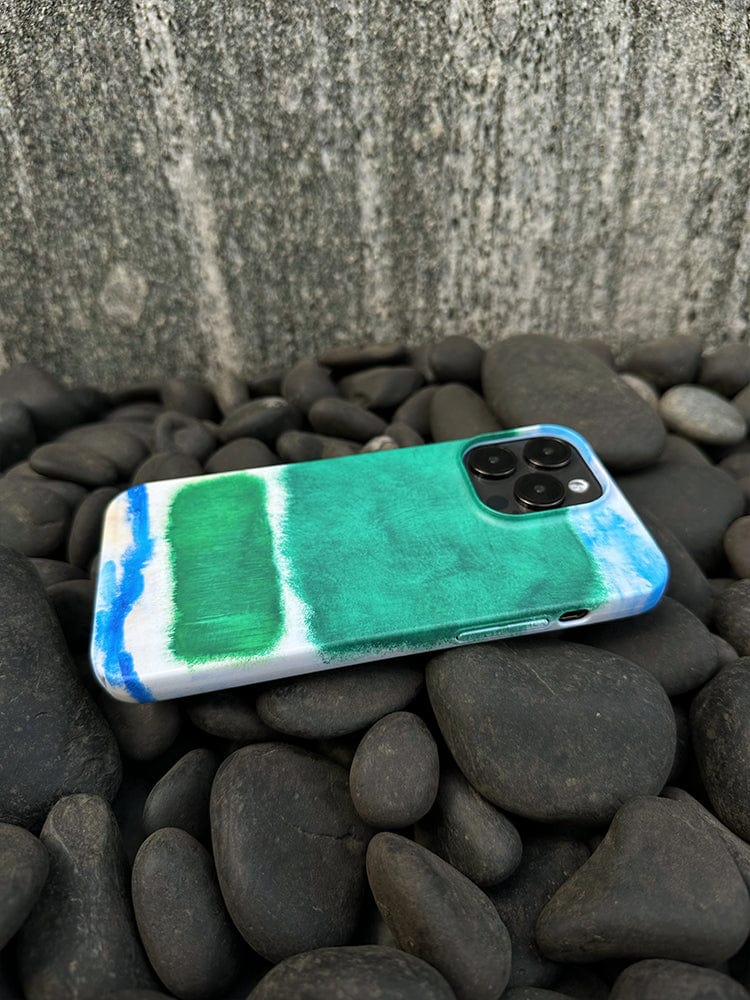 An iPhone 16 Pro Max with an Abstract Color Block case, featuring a modern art-inspired, minimalist design in green and blue, lies face down on a bed of black pebbles. The phone, boasting a triple-camera setup on the back, is set against the backdrop of a textured gray wall.