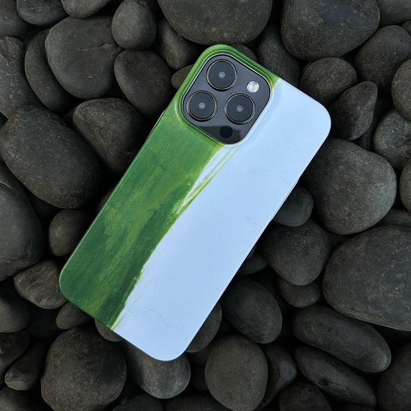 A smartphone adorned with a Minimalist Green Brushstroke iPhone 16 Pro Max case, featuring an artistic abstract design, placed on a bed of smooth, dark pebbles. Only the back of the phone is visible, showcasing the camera lenses and highlighting the elegant color contrast of the ultra-slim case.