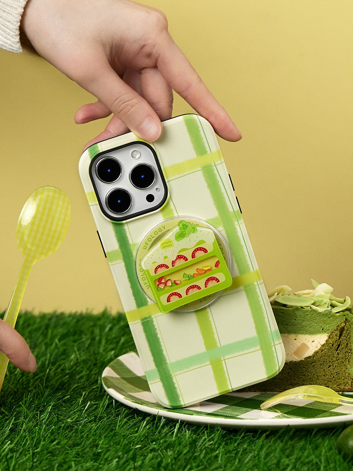 Hand holding a Fresh Picnic MagSafe iPhone 16 Pro Max Case with a green plaid design, featuring a sandwich-themed stand. Nearby, a spoon and a slice of matcha cake are placed on artificial grass.