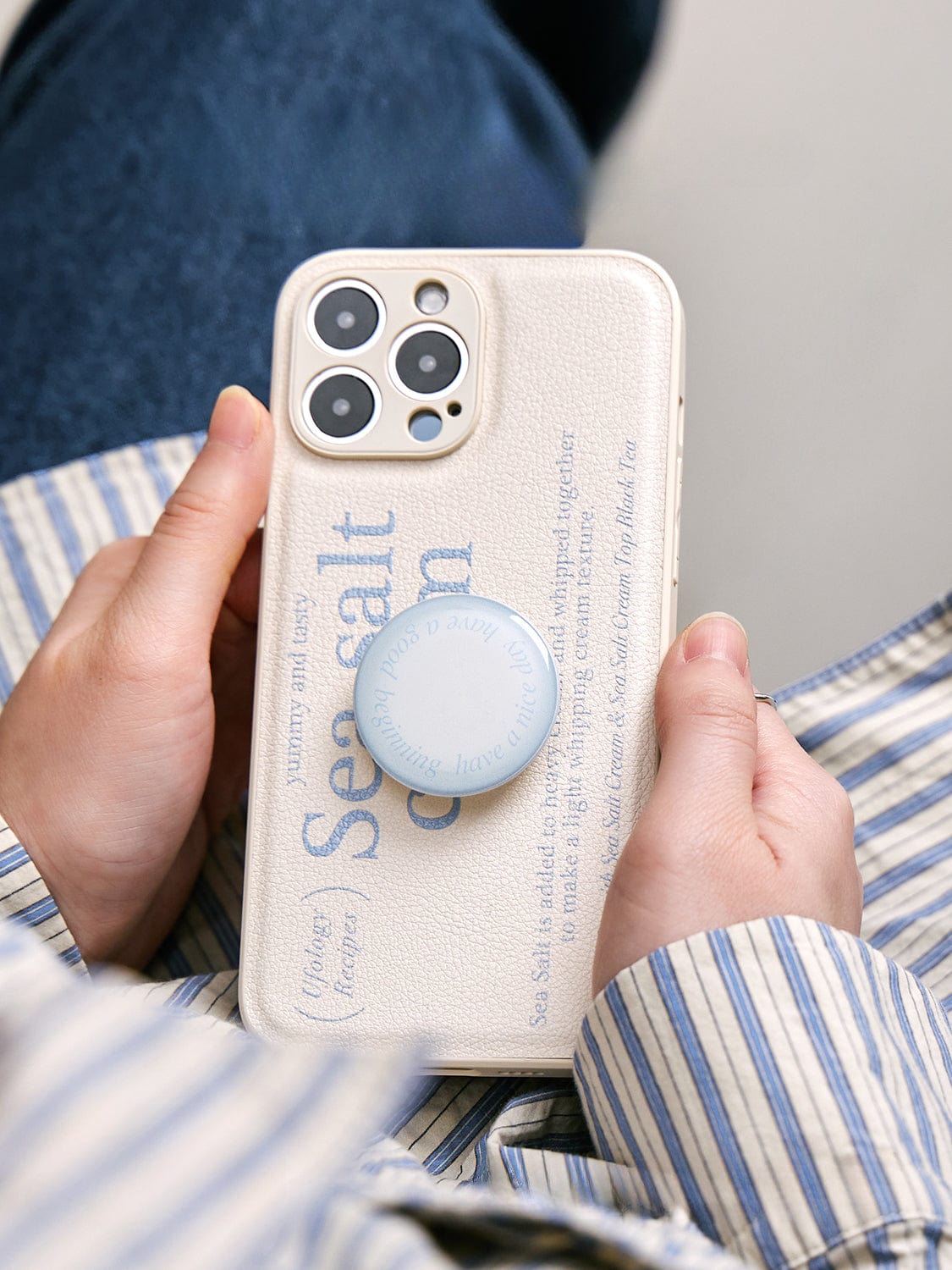 A person holds a Sea Salt Cream MagSafe iPhone 16 Pro Max case with a minimalist textured design, featuring text and a blue pop socket attached to it.