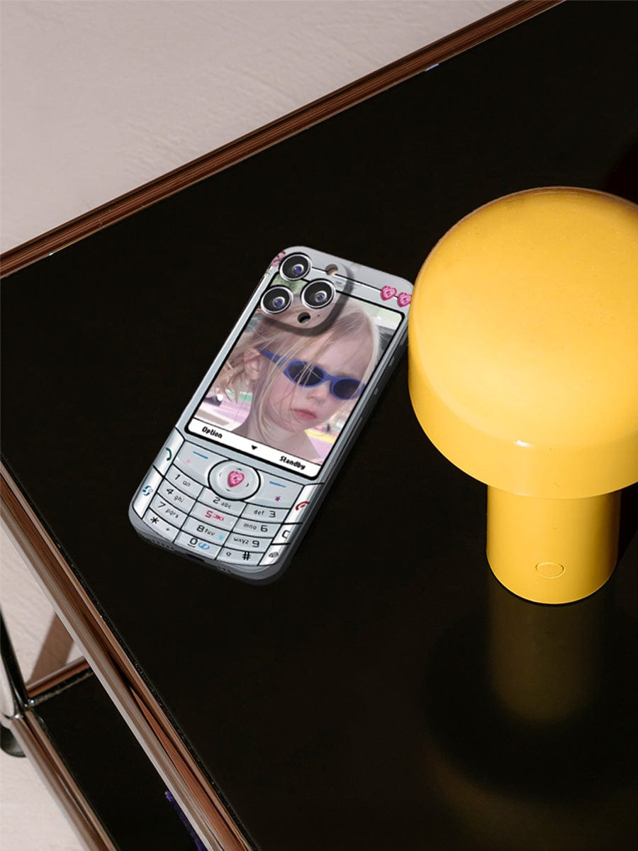 A smartphone, adorned with the Retro Flip Phone iPhone 16 Pro Max Case featuring a Y2K nostalgia design, is placed on a dark reflective surface next to a yellow cylindrical lamp. The phone screen displays an image of a child wearing sunglasses.