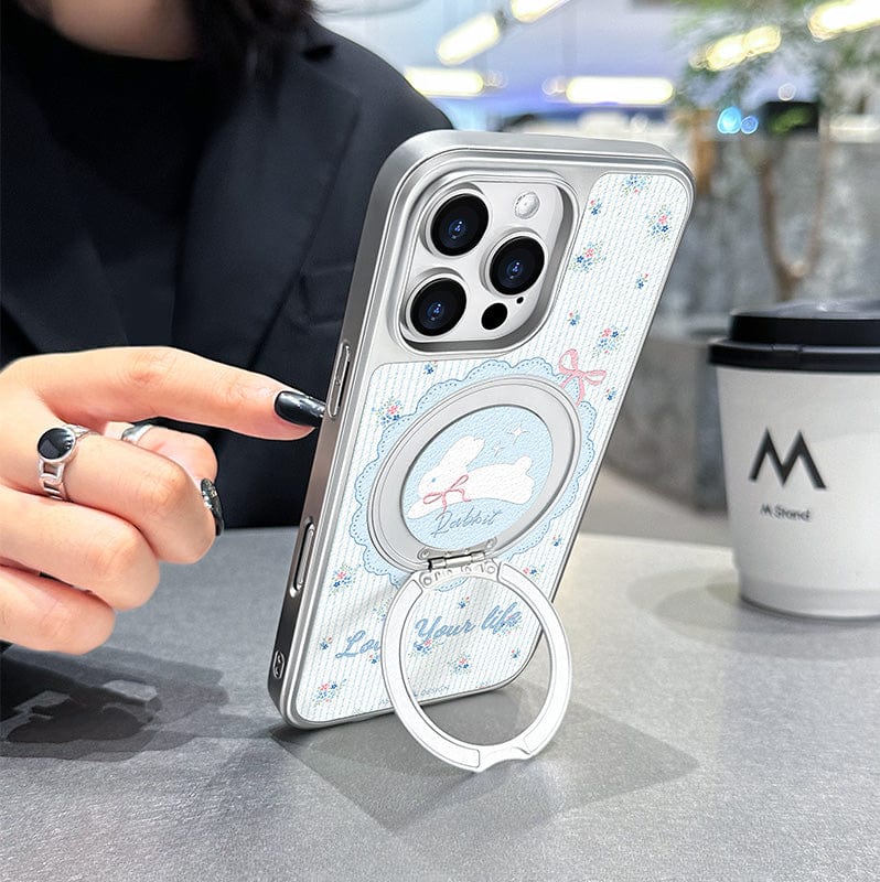 Close-up of a person holding an iPhone 16 Pro Max with a Cute Case featuring a floral rabbit design. A coffee cup sits on the table in the background.