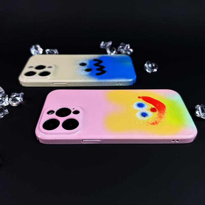 Two iPhones lying flat against a black background. The phone in the foreground has a pink case with a colorful, abstract, smiley face design. The Colorful Spray Art iPhone 16 Pro Max Case Set - Playful Faces in Vibrant Graffiti Style in the background is white with a blue and black design resembling eyes and a mouth. Small crystals are scattered nearby.