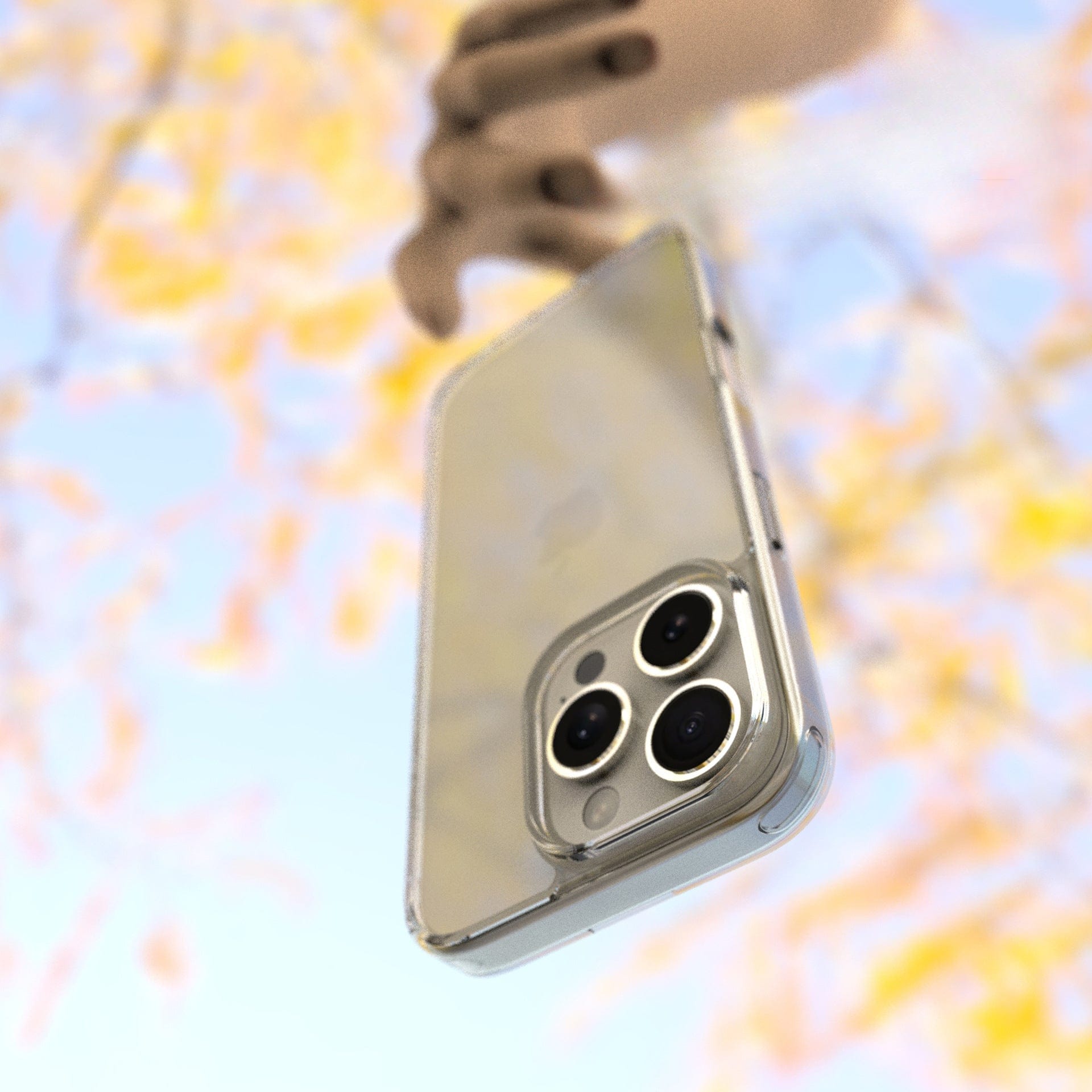 A hand holding an iPhone 16 Pro Max encased in an Ultra-Thin Clear Case with MagSafe, featuring a triple camera setup and shockproof bumper corners, against a blurred background of yellow and pink flowers.