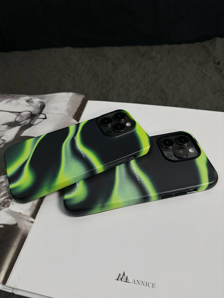 Two smartphones with green and black gradient cases lie on an open magazine displaying monochrome images. The phones, featuring the sophistication of the Abstract Wave iPhone 16 Pro Max Case - Sleek Design with "Happier Than Ever" quote, are placed next to each other, showcasing their rear cameras and stylish designs.