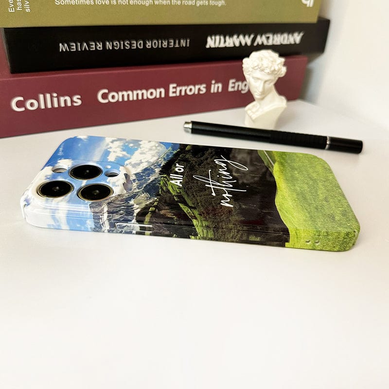 A smartphone encased in a Mountain Landscape iPhone 16 Pro Max Case - Inspiring Scenic Design with "All or Nothing" Quote is placed on an open shelf, leaning against a stack of books titled "Common Errors in English," "Meditations," and "Anthology." A pen and a small bust sculpture are also on the shelf.