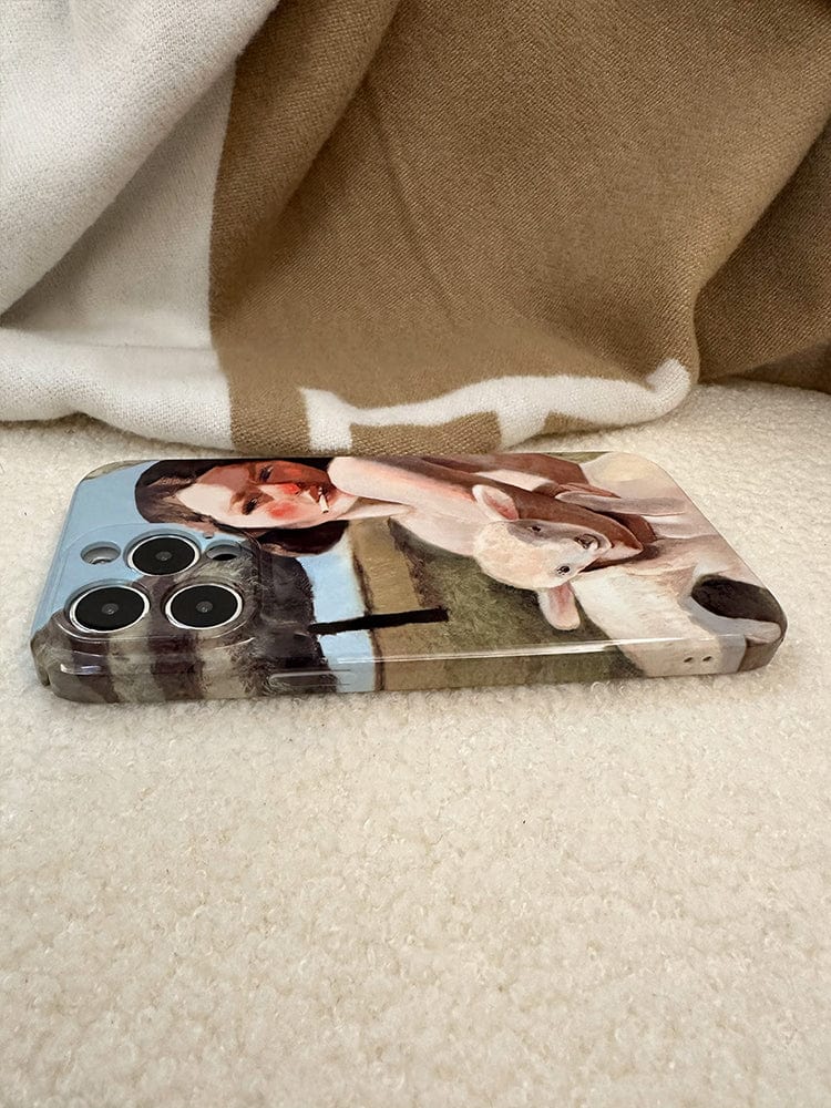 A smartphone enclosed in a "Pastoral Art iPhone 16 Pro Max Case - Vintage-Inspired Design with a Modern Twist" featuring a painting of a woman in a white dress holding a lamb. The phone is resting on a soft, beige and white blanket, with the camera lenses facing upwards.