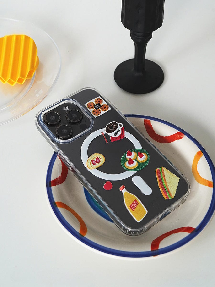 Brunch Time MagSafe iPhone 16 Pro Max Case with a cute 3D food design, featuring colorful stickers of a sandwich, rice bowl, condiments, and sushi displayed on a white plate with a blue and orange rim. Nearby are a yellow plastic grid piece, a black cup, and part of a translucent lid.