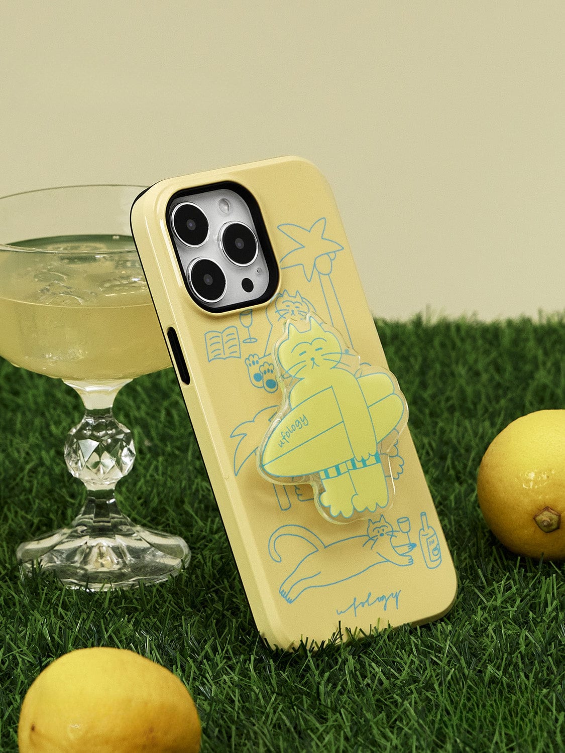 The Tropical Cat MagSafe iPhone 16 Pro Max Case in sunny yellow, featuring a relaxed cat with a book, stands upright on artificial grass. Beside it sits a crystal glass filled with a light-colored drink, accompanied by two lemons.