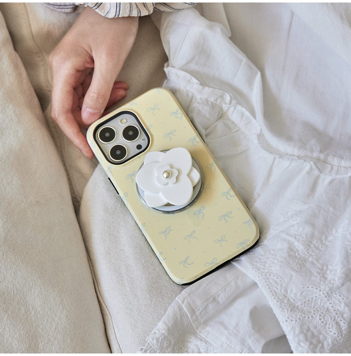 A hand resting on white fabric holds a smartphone with an Elegant Blossom MagSafe iPhone 16 Pro Max Case, featuring a chic floral design and a pearl flower stand.