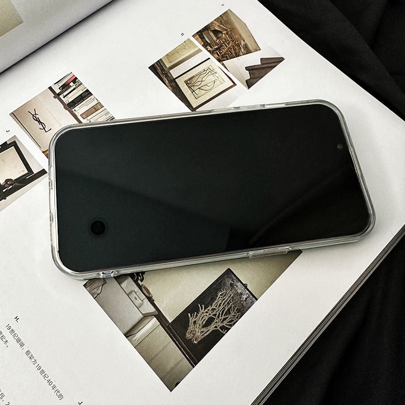 A Snaccident iPhone 16 Pro Max Case - Humorous Vintage Art Design with Full Protection sits beside a smartphone with a black screen, which lies face-up on an open magazine showcasing various interior design photos. The pages depict modern home decorations and furniture in neutral tones, all resting on a black fabric surface.