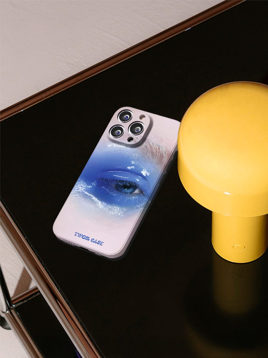 A Dreamy Blue Eye iPhone 16 Pro Max Case, featuring a surreal art design of a blue eye and wave, is placed on a sleek, dark surface next to a small, bright yellow cylindrical object. The smartphone's camera lenses are visible at the top of the elegant all-inclusive protective cover.