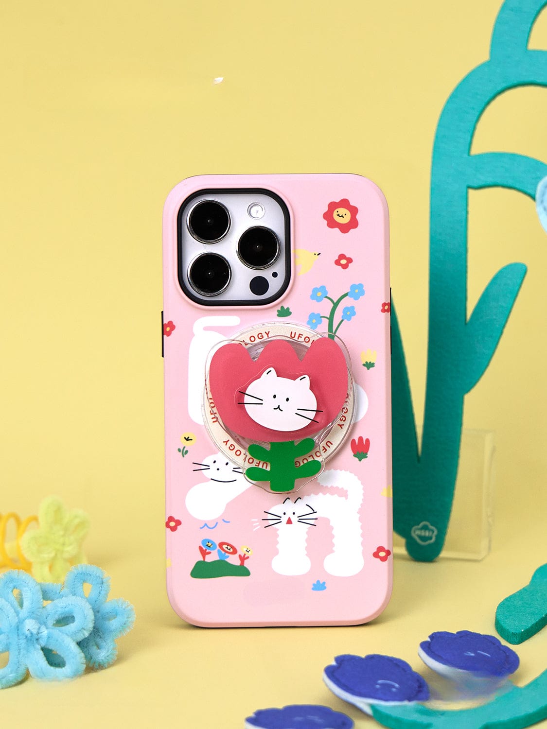 A Spring Kitty MagSafe iPhone 16 Pro Max Case, showcasing a playful floral design and featuring a built-in cat stand, is propped up vertically against a light yellow background with colorful decorations.