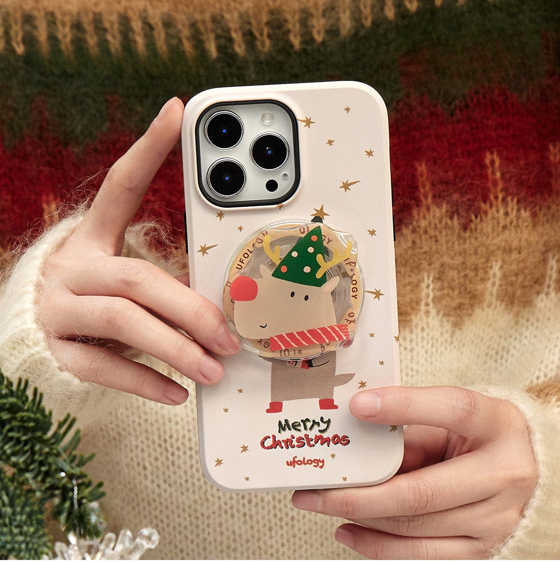 Close-up of a person's hands holding an iPhone 16 Pro Max with the Rudolph Joy MagSafe Case, which features a festive reindeer design and the text "Merry Christmas." The background includes a festive tree.