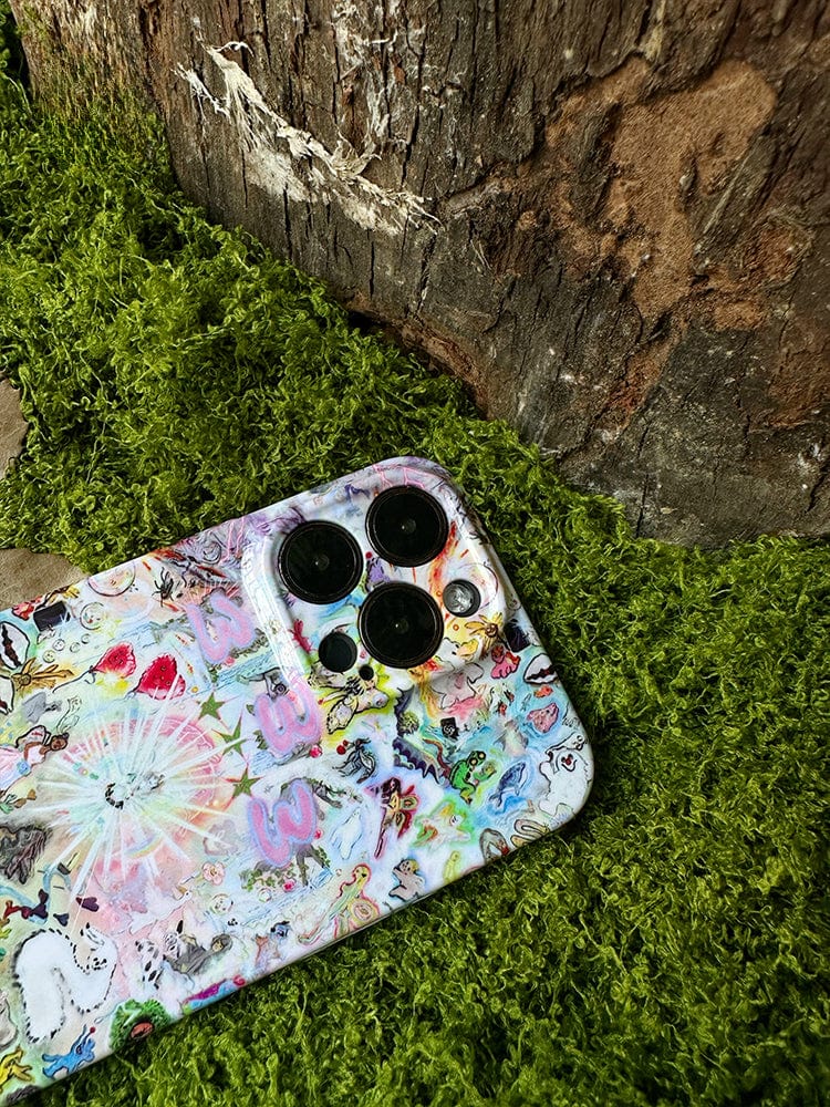 A Whimsical Dreamscape iPhone 16 Pro Max, adorned with a fantasy-inspired artistic case featuring intricate and colorful designs, lies on a patch of green moss. The phone is positioned near a large, textured tree trunk in an outdoor setting.