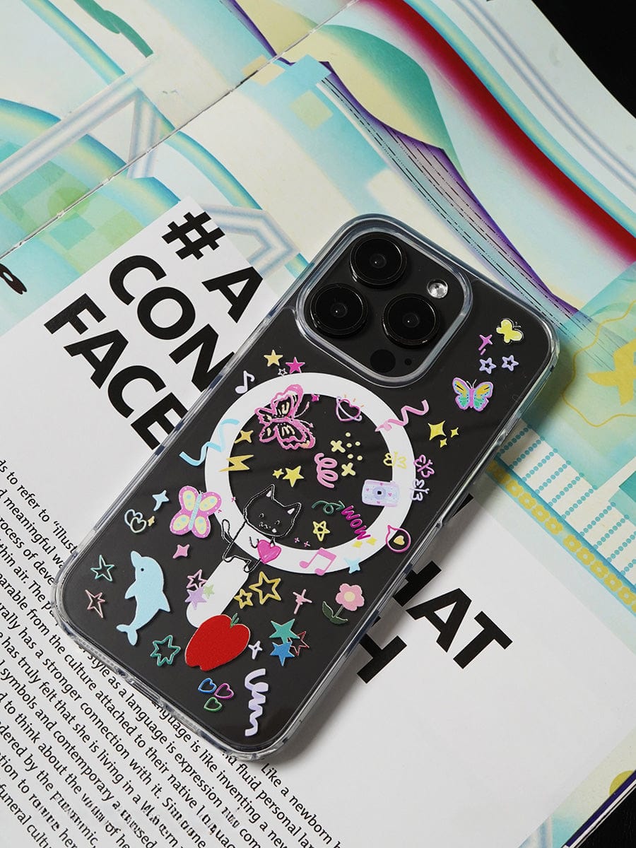 A smartphone, specifically an iPhone 16 Pro Max housed in the Whimsical Dreams MagSafe Case — a transparent protective cover featuring a playful design with colorful stickers of stars, dolphins, and butterflies — is placed on an open magazine.