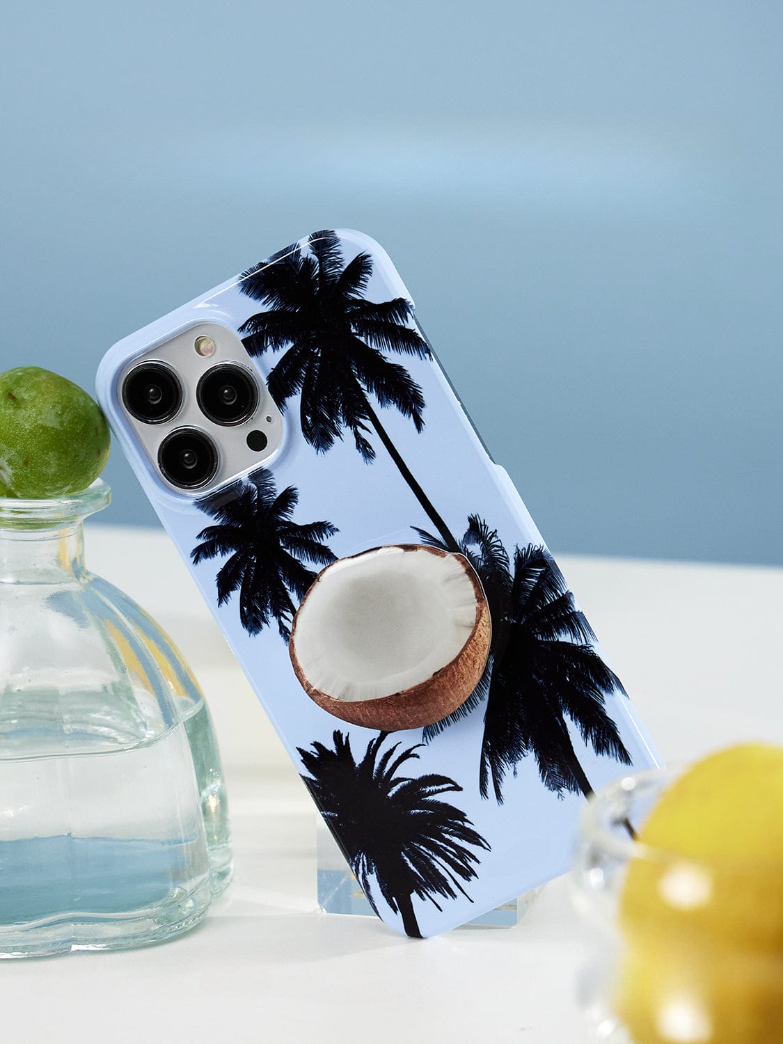 The Tropical Escape MagSafe iPhone 16 Pro Max Case, featuring a palm tree design with black silhouettes and a 3D coconut shell on a light blue background, is propped up against a clear glass vase containing a green lime. Blurred objects are visible in the foreground and background.