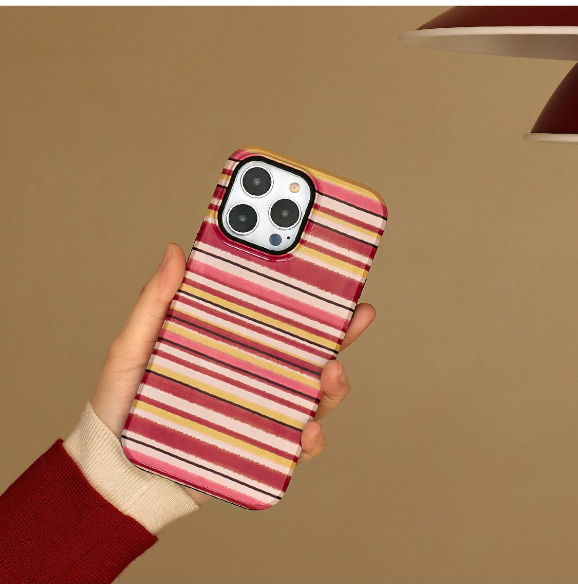 A hand is holding an iPhone 16 Pro Max encased in the Autumn Stripes MagSafe Case featuring a cozy mug design with pink, white, and yellow stripes. The phone is set against a neutral beige background. The person's sleeve is red, and the iPhone is tilted slightly to showcase both the case design and the phone's camera.