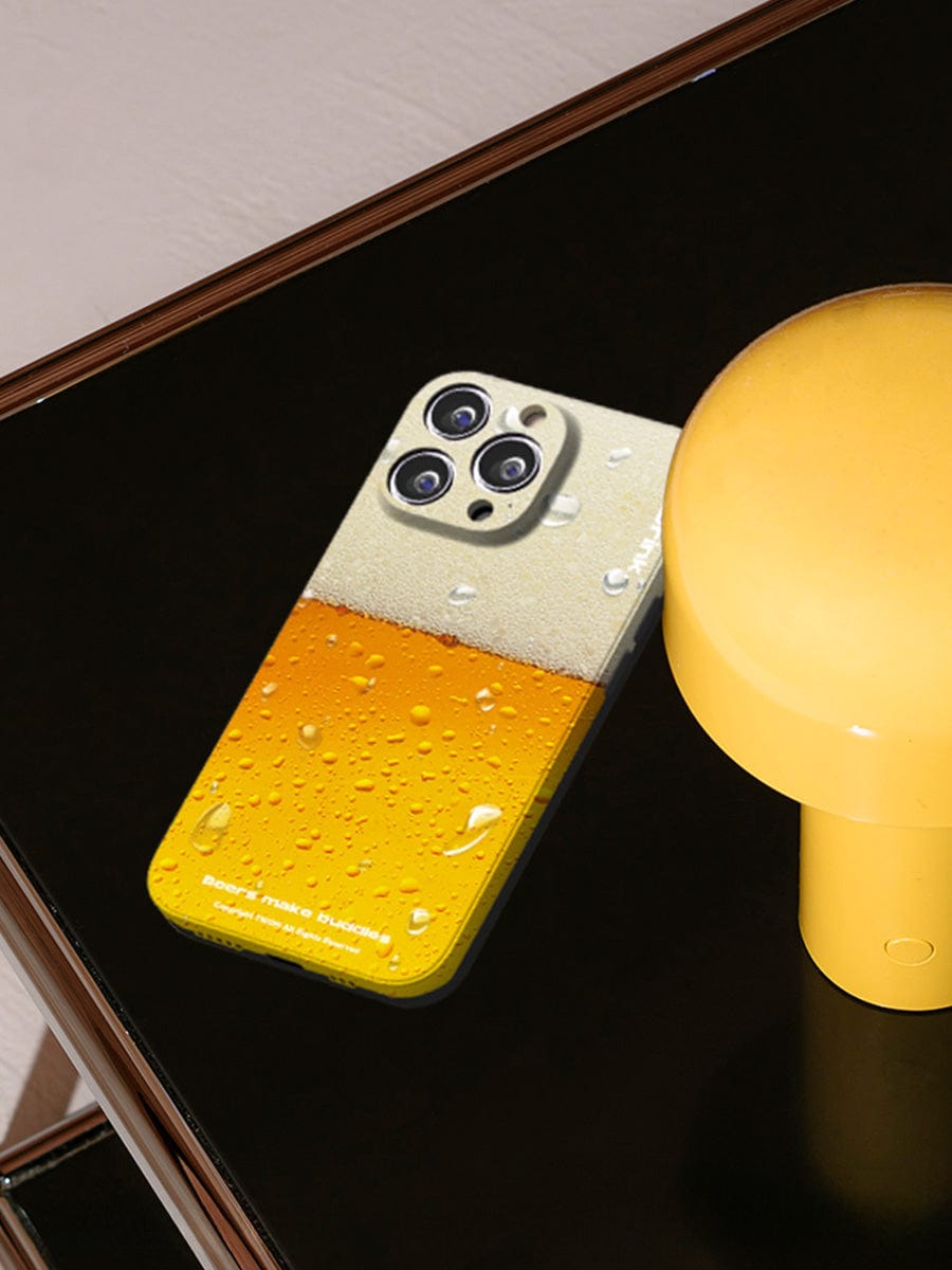 A Beer Buddies iPhone 16 Pro Max Case, featuring a fun beverage design with frothy beer and condensation droplets, rests on a black surface near a yellow lamp.