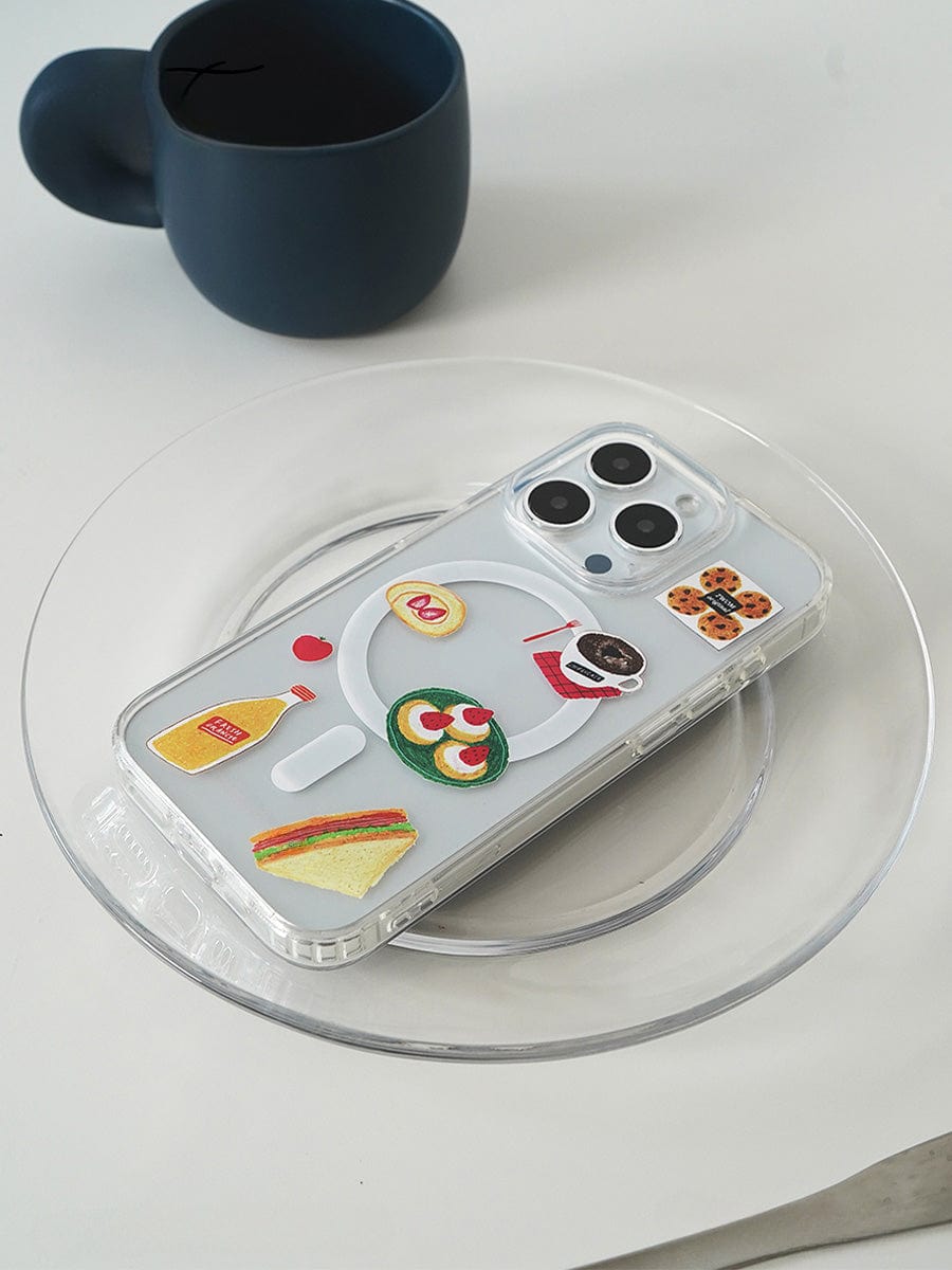 The Brunch Time MagSafe iPhone 16 Pro Max Case, adorned with fun 3D food designs like a sandwich, noodle bowl, and avocado toast, showcases its protective cover on a silver smartphone. The phone lies on a clear glass plate with a dark-colored cup in the background on a white surface.