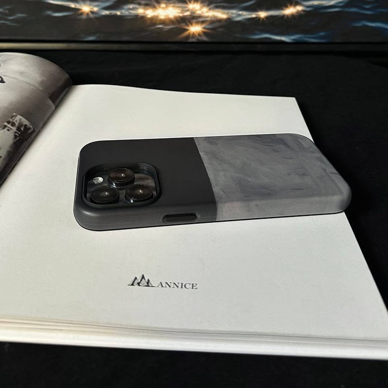 A sleek, minimalist iPhone 16 Pro Max case in elegant dark green and gray is placed on an open book lying on a black surface. The book features a small illustration of mountains and the word "ANNICE" printed on one of its pages. In the background, twinkling lights add a touch of sparkle.