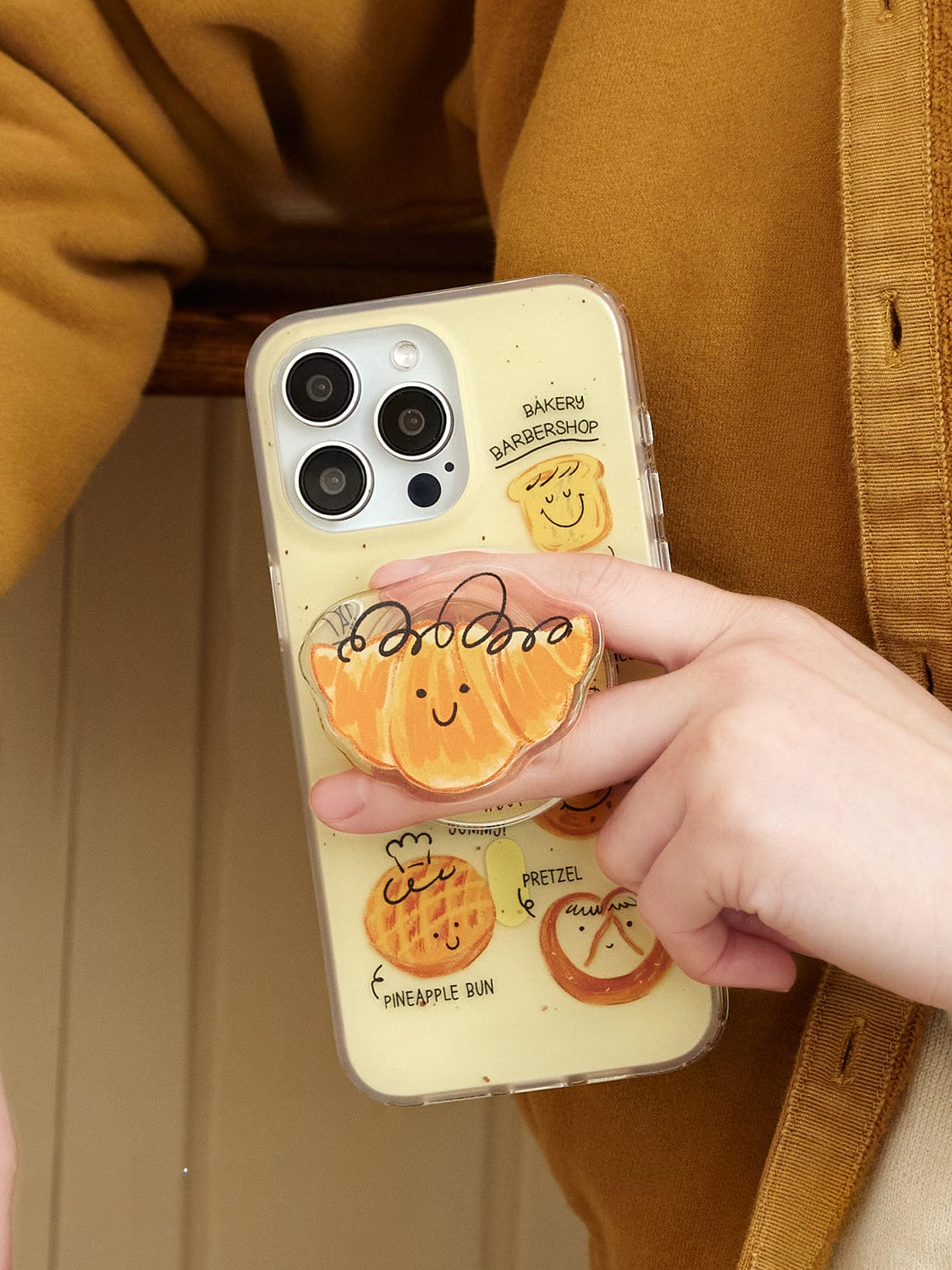 A person is holding the Bakery Friends MagSafe iPhone 16 Pro Max Case, which showcases a cute bread design with colorful, cartoonish illustrations of baked goods like pineapple buns and pretzels. One illustration highlights a smiling orange bread bun with the word "baker" above it.