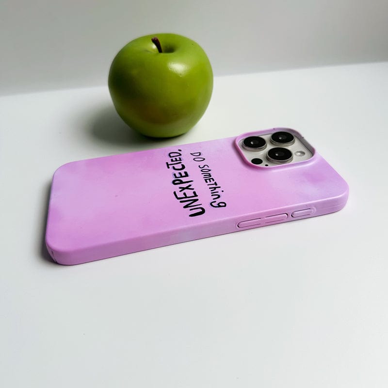 A green apple sits on a white surface next to an Inspirational Pink iPhone 16 Pro Max Case featuring the text, "UNEXPECTED. Do something." The phone case boasts a 'Do Something Unexpected' quote design and offers ultra-slim, durable protection.