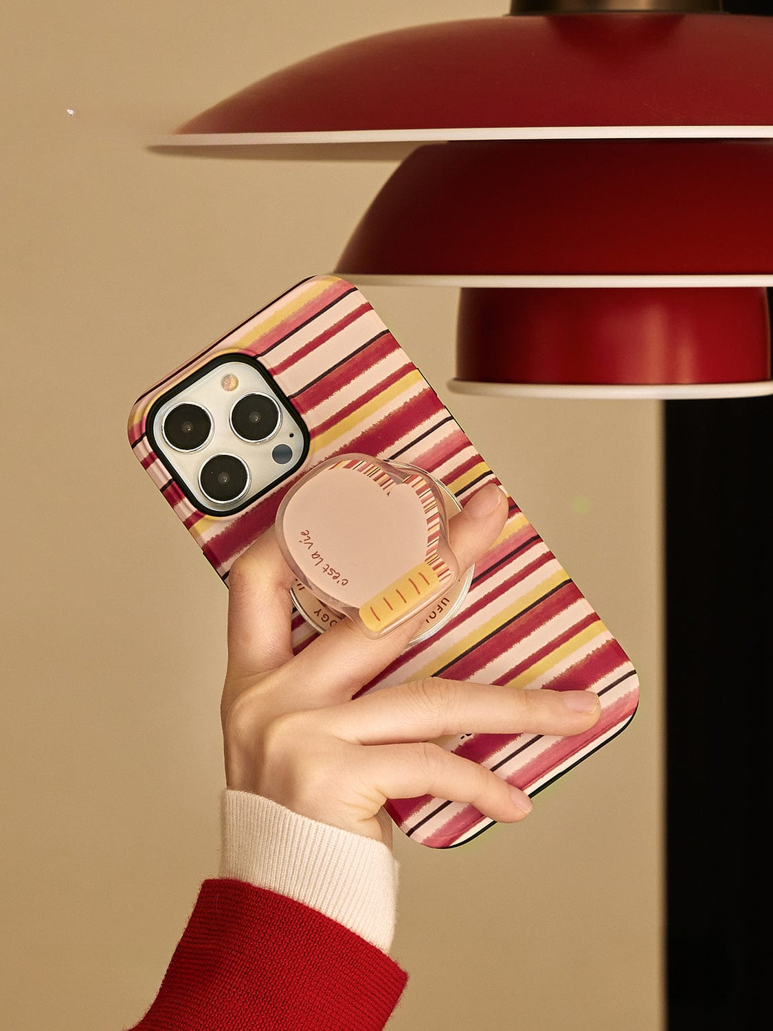 A hand holds an iPhone with the Autumn Stripes MagSafe iPhone 16 Pro Max Case, featuring horizontal stripes in red, yellow, and white and a cozy mug design grip stand. Above the phone, a red lamp with a white interior casts light on the scene.