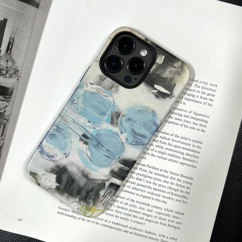 An iPhone 16 Pro Max with a multi-lens camera lies on an open book. The phone features an Abstract Blue Circles case, showcasing a modern art-inspired design with blue and white patterns resembling paint strokes. The text on the book page is partially visible but not readable.