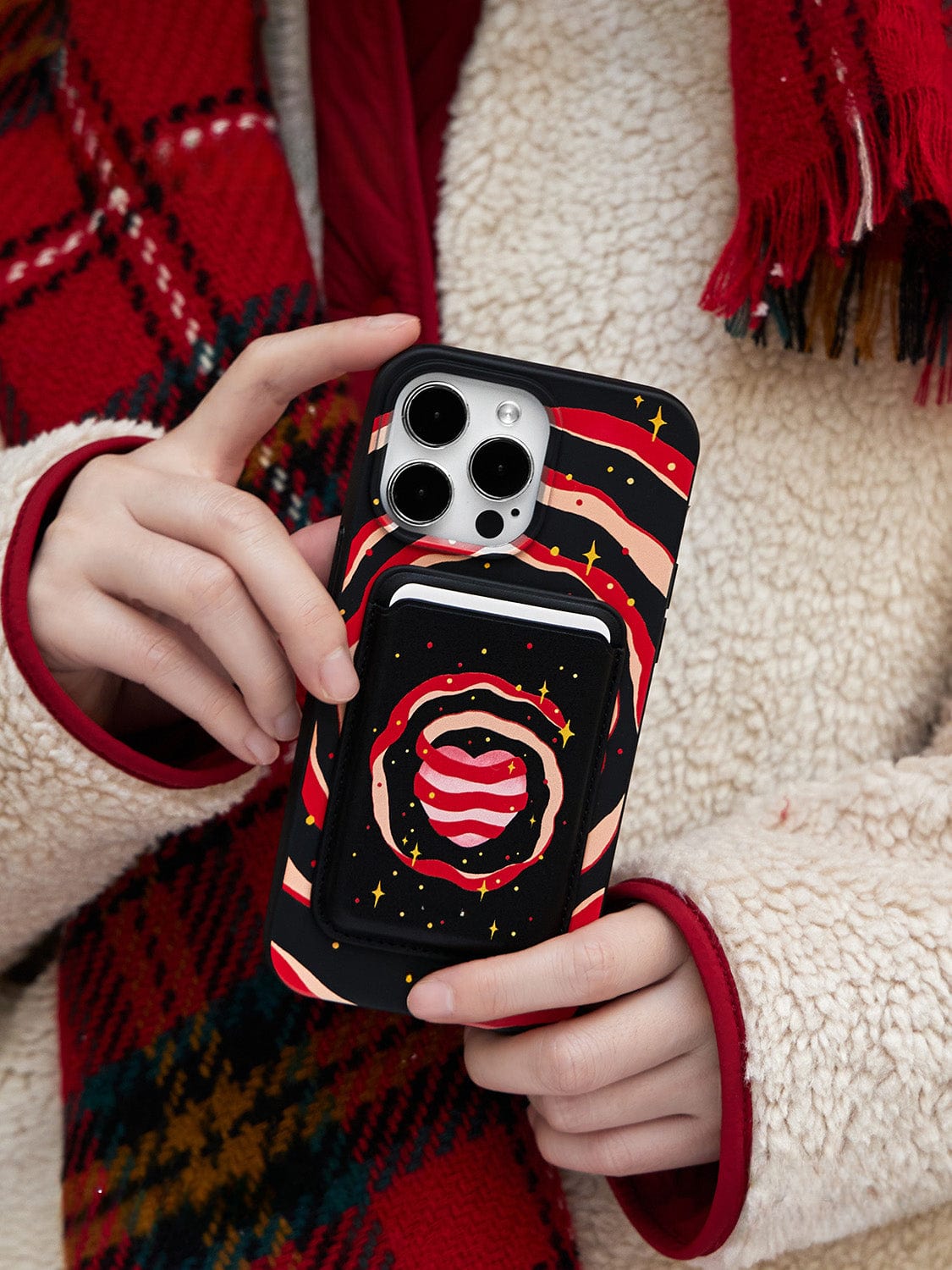 A person wearing a red plaid scarf and a light-colored, fuzzy jacket holds a smartphone with the Galaxy Heart MagSafe iPhone 16 Pro Max Case. The case displays a vibrant, cosmic pattern of red and pink swirls with yellow stars and includes a matching pocket accessory.