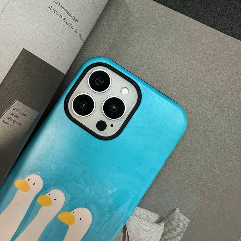 An iPhone 16 Pro Max with a Whimsical Three Ducks Case in playful blue and yellow design is placed on an open magazine. The phone's cameras are visible, and the magazine pages are in grayscale.