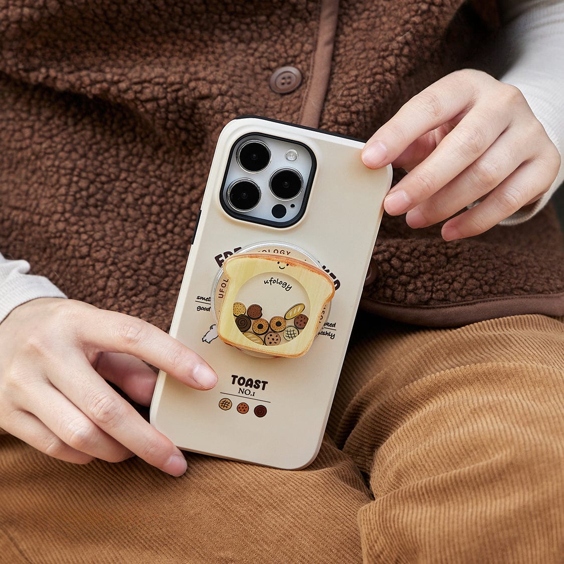 A person dressed in a brown fleece jacket and brown corduroy pants holds an iPhone 16 Pro Max encased in the Morning Toast MagSafe Case, featuring a cute breakfast design with cartoon toast and a bear. The hand holding the phone is wearing a white shirt beneath the jacket, and the person is sitting down.