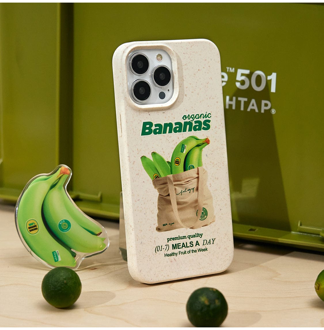 An Organic Bananas MagSafe iPhone 16 Pro Max Case, featuring a fun fruit design with a banana stand, rests against a closed green storage container labeled "FOLDING CONTAINER 501 LIGHTTAP." In front, a banana-shaped keychain and small green fruits are displayed.