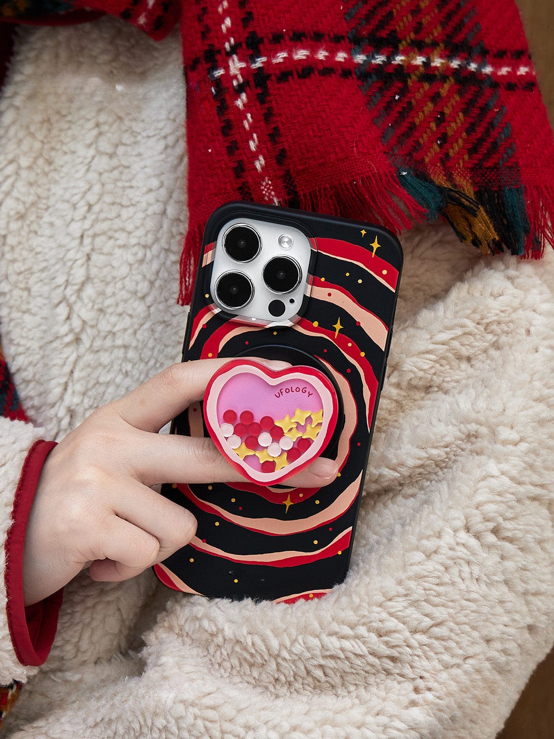 A person wearing a red plaid scarf and a beige, fluffy jacket holds a smartphone in one hand. The Galaxy Heart MagSafe iPhone 16 Pro Max Case is black with wavy red and white stripes, featuring a heart-shaped accessory filled with small multicolored decorations.