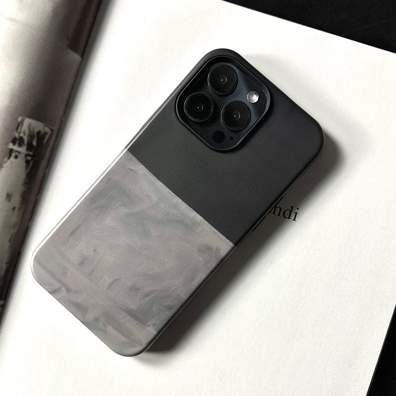 A smartphone encased in a Minimalist Two-Tone iPhone 16 Pro Max Case, featuring an elegant design with a dark green upper half and a translucent gray lower half, is placed face-down on an open book with visible text. The case houses three lenses in its camera setup.