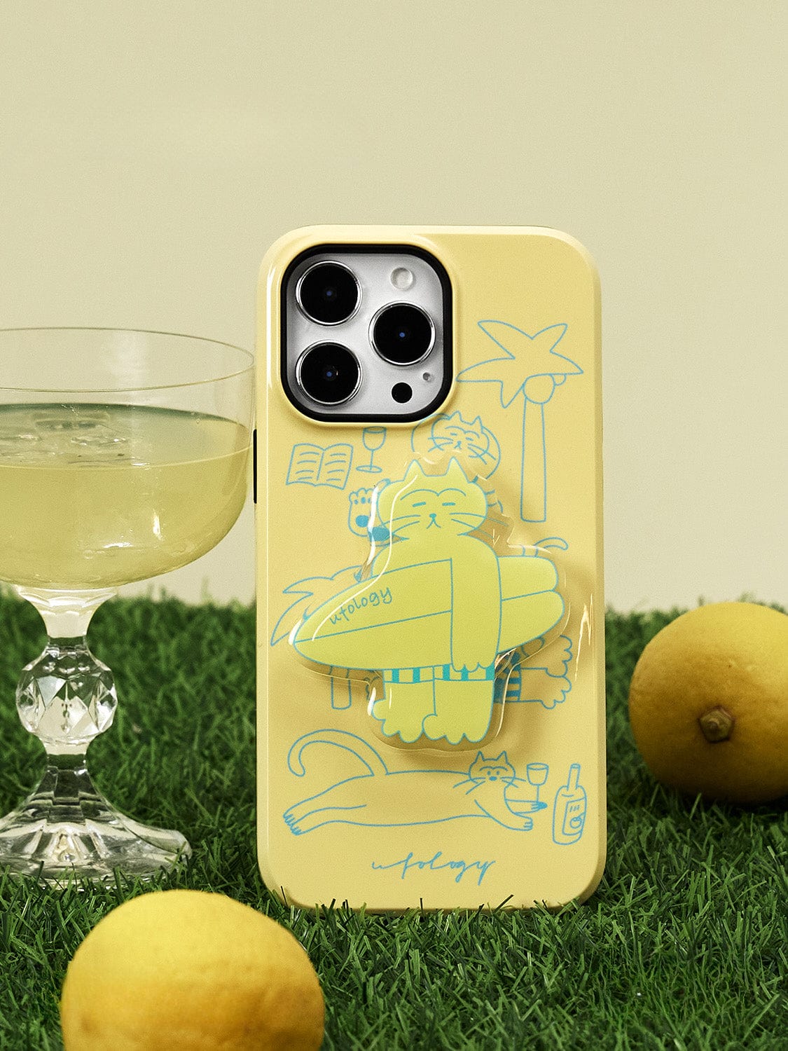 The Tropical Cat MagSafe iPhone 16 Pro Max Case, featuring a sunny yellow design with a relaxed cat stand amidst line art of a drink, book, and surfboard, is placed on artificial grass next to a crystal glass of a light beverage. Two lemons are also visible near the phone case.