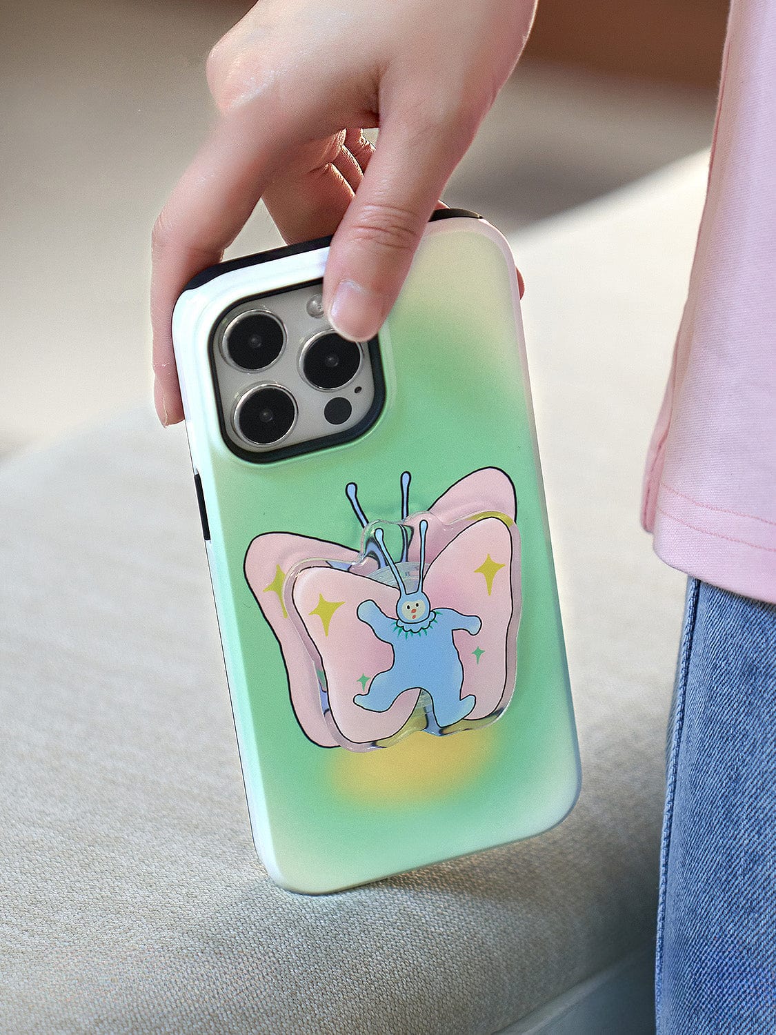 A hand holding an iPhone 16 Pro Max with a Golden Sunset MagSafe case, featuring an elegant gradient design and a ring stand. The gradient background transitions from green to light blue. The person is wearing a light pink shirt and blue jeans.