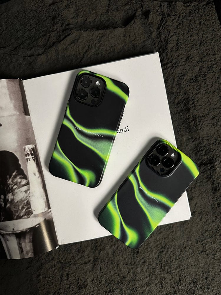 Two smartphones with matching Abstract Wave iPhone 16 Pro Max cases, featuring a sleek design and the "Happier Than Ever" quote, rest on top of an open book. The phones are placed on a textured, dark-colored surface. One phone is slightly larger than the other. The book and the background provide a contrasting backdrop.