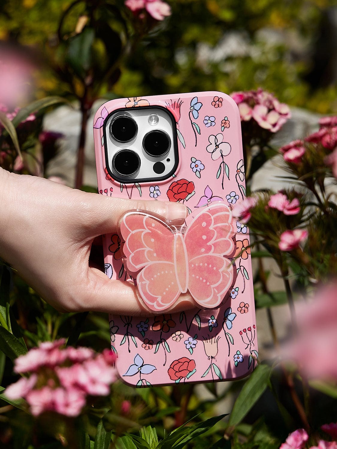 Hand holding a Blossom Butterfly MagSafe iPhone 16 Pro Max Case with a floral pink design and butterfly stand among pink flowers.