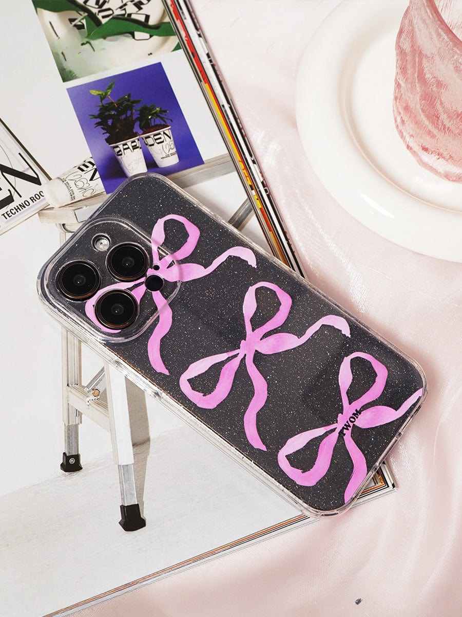 A smartphone with a Pink Ribbon Bow Transparent iPhone 16 Pro Max Case, showcasing four large pink bows in a sparkling clear design, is placed on a magazine. The magazine displays an image of a plant on a white ladder. A portion of a pink object, potentially fabric or an accessory, is visible on the right side.