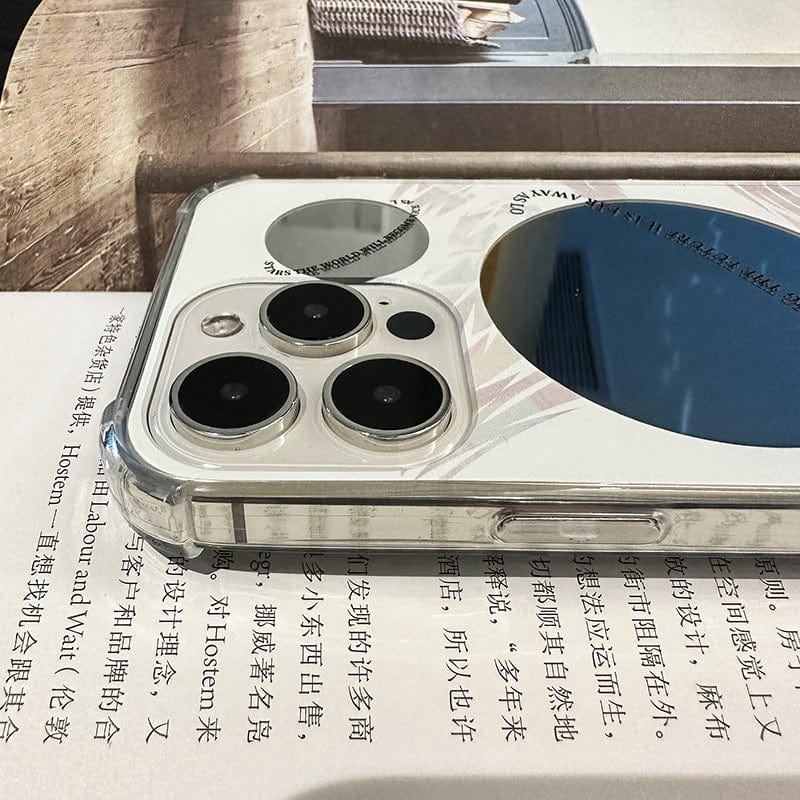 A close-up of the Futuristic Distant Planet iPhone 16 Pro Max Case - Sleek and Protective, showcasing its transparent design and a distinctive round decorative element adjacent to the camera lenses. The phone is positioned on what seems to be a book or magazine with Chinese text. The scene also includes a wooden surface and a partially visible item in the background.