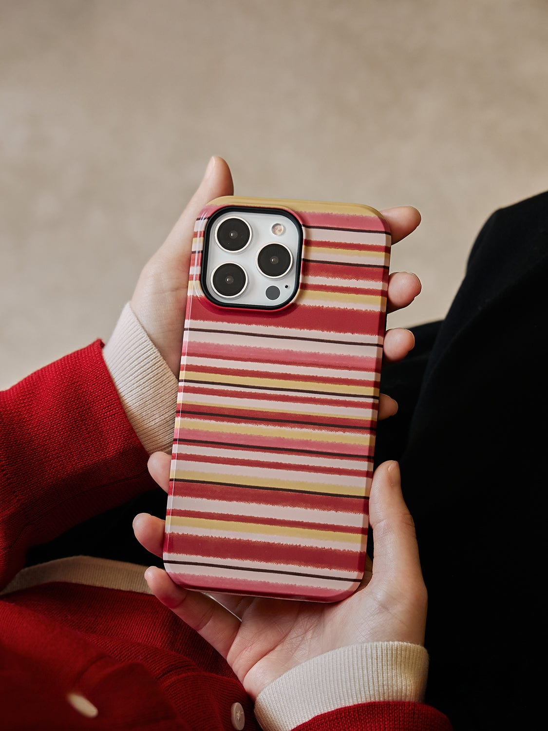 A person wearing a red sweater is holding an iPhone 16 Pro Max with the Autumn Stripes MagSafe Case, which features a cozy mug design and red, pink, and white stripes. The background is blurred and appears to be indoors.